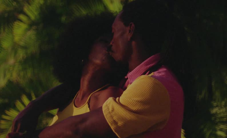 Side view of female dancer with curly hair and yellow dress and male dancer with braids and pink yellow shirt standing in front of trees on grass making out at night