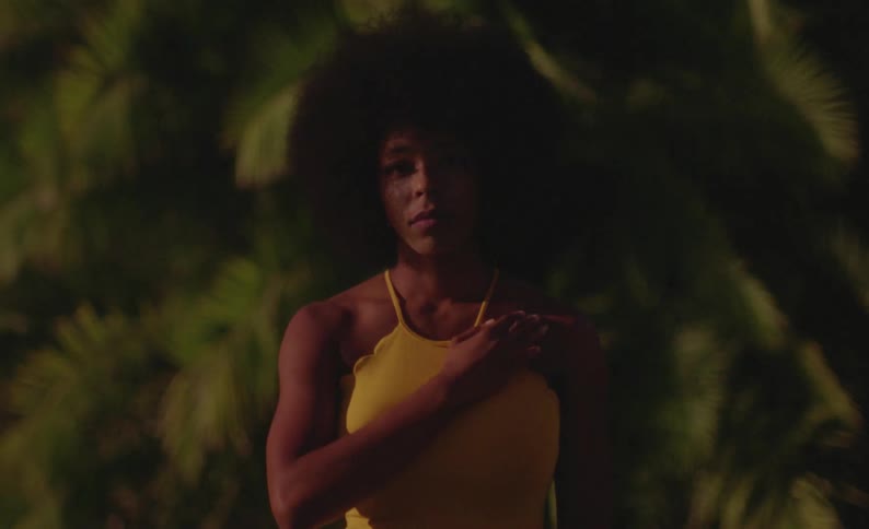 Black woman in front of a background of green leaves wearing a yellow outfit and putting her hand on her chest
