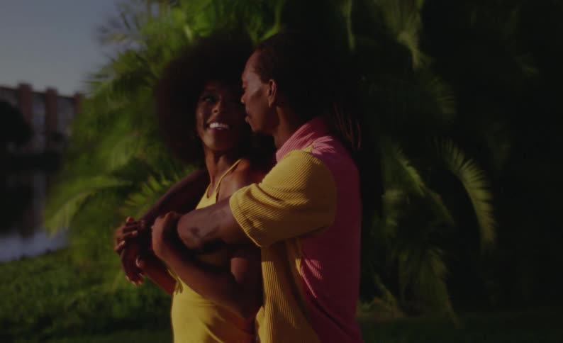 Man back hugs woman and they smile at each other as they stand outdoors near pond