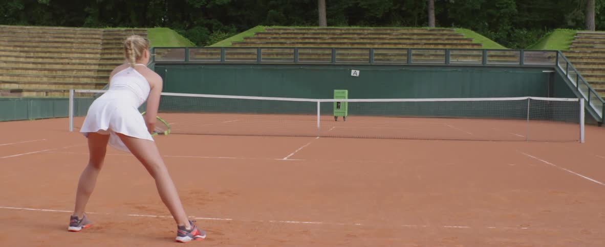 A woman readies herself as she stands across from a tennis ball machine on an outdoor tennis court during the day
