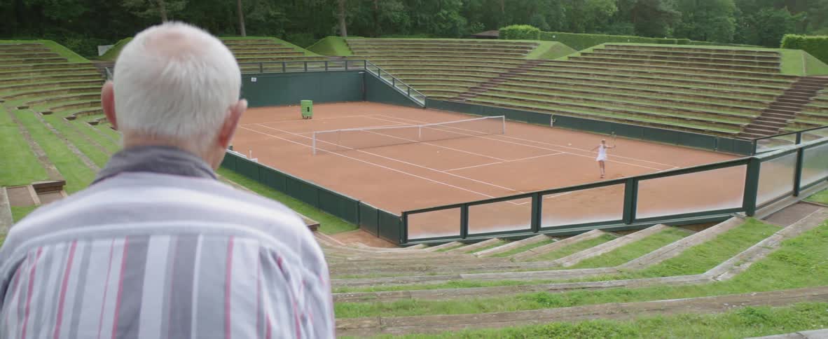 Elderly man with gray hair and gray shirt watches female tennis player playing tennis in tennis court