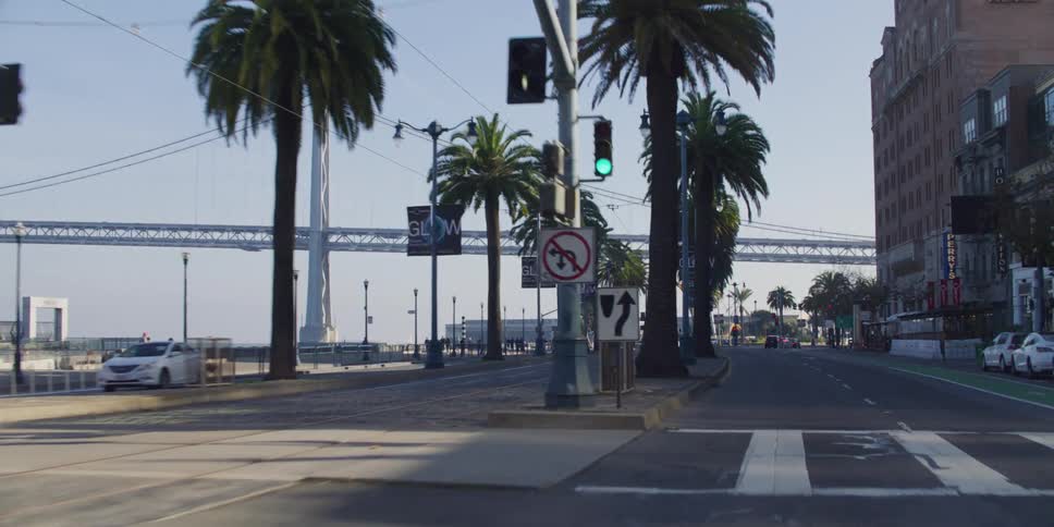 Cars move through intersection passing pedestrians and buildings as they head toward suspension bridge