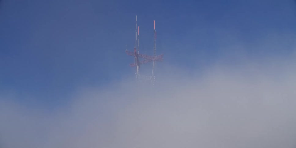 Antennas atop a communication tower peeking over white clouds on the blue sky