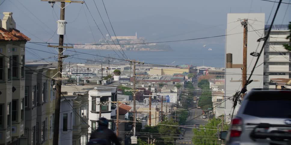 Hilly city street with poles and vehicles overlooking sea with small island