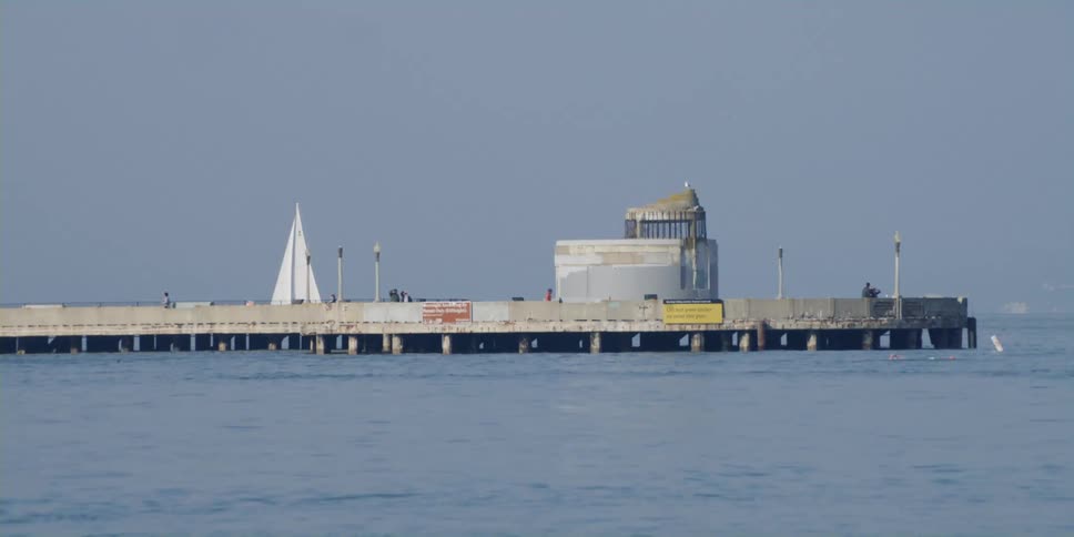 A sailboat sails past a pier where people are walking and jogging and swimming near by