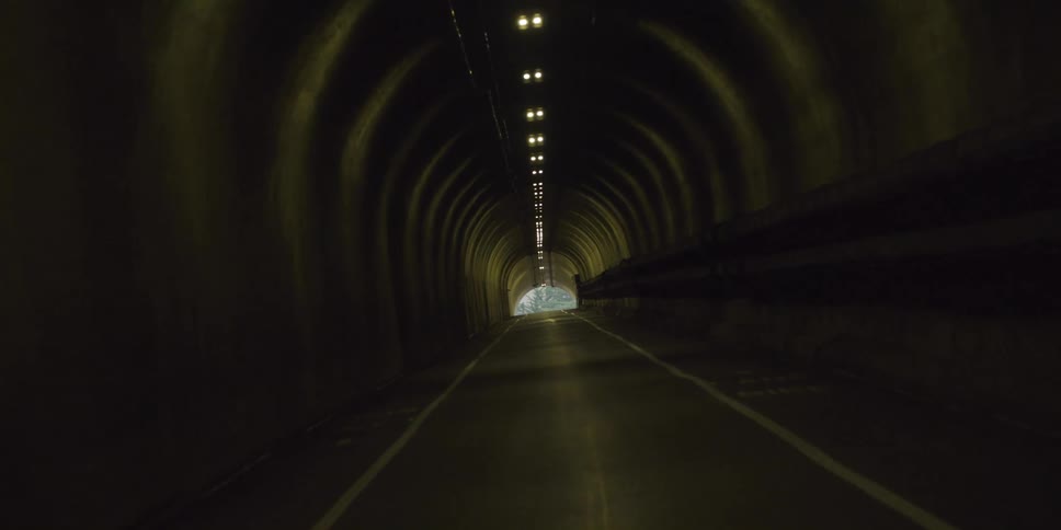 A road through a dark tunnel with lights on the ceiling and trees and sunlight at the end of the tunnel