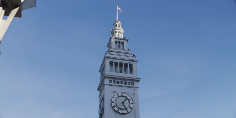 American flag flutters on top of clock tower standing over streetlamp