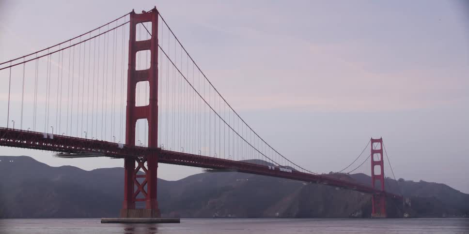 A suspension bridge is stretched across water with mountains in the distance