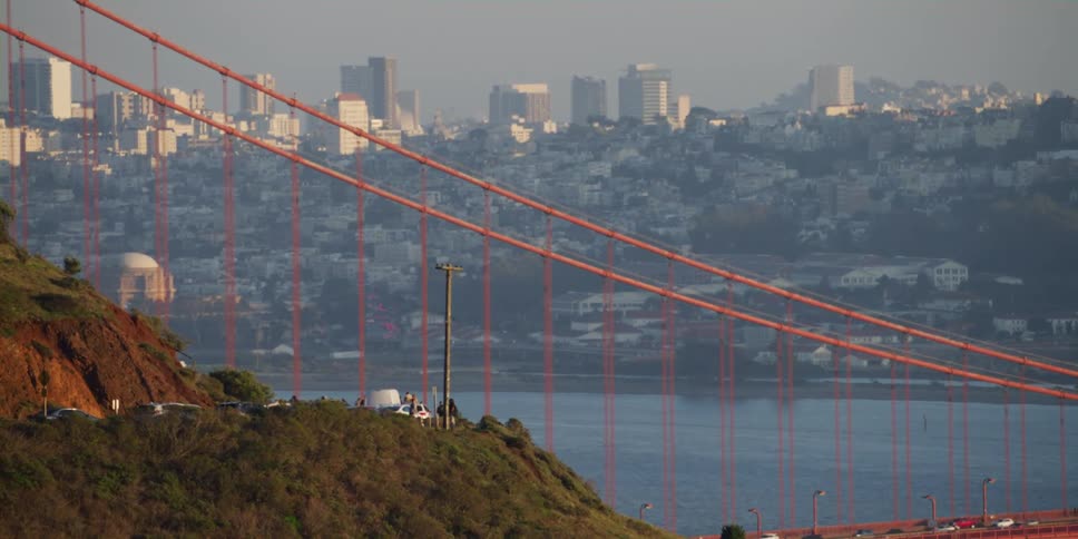 Cars drive across a bridge in front of a city skyline while people walk on the hill above