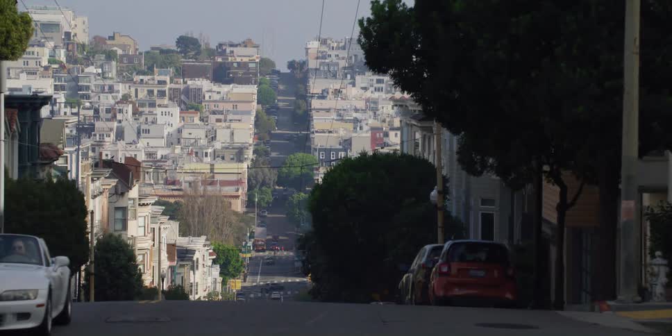 A car drives up a neighborhood street lined with cars as a person walks on the sidewalk