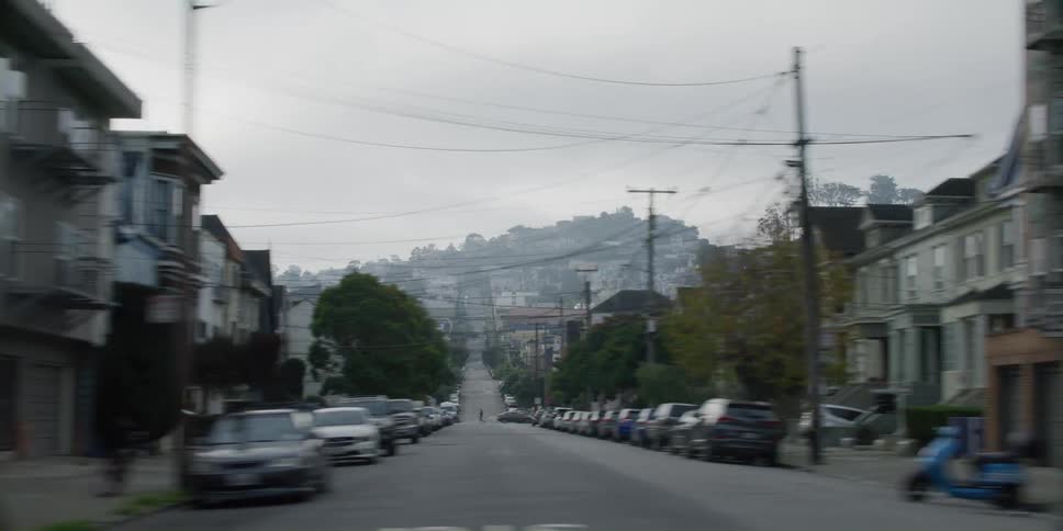 Fog covers buildings on hills at end of neighborhood street lined with parked vehicles