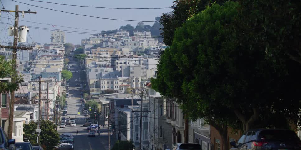 Man walks along sidewalk near top of hill carrying a lamp while traffic passes through intersection at bottom of hill