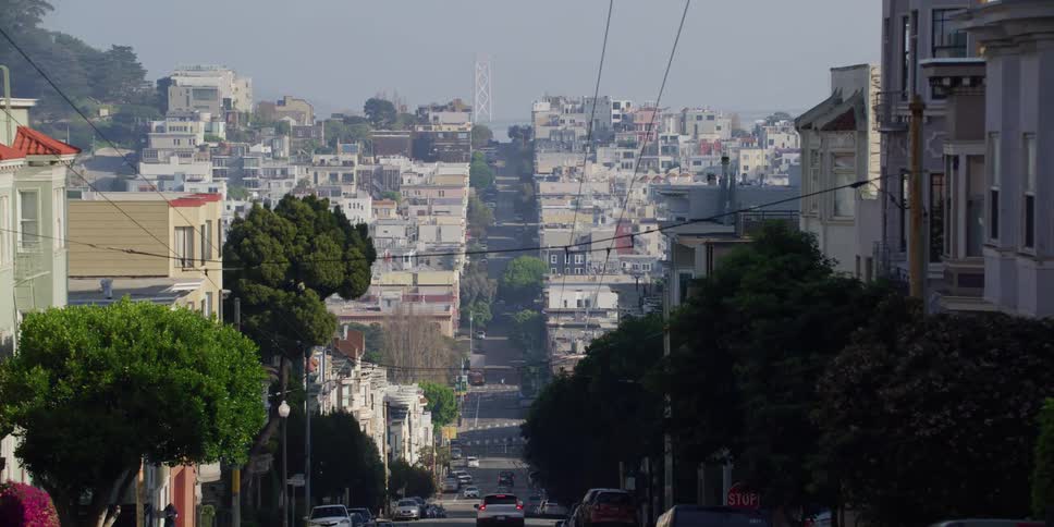 Vehicles drive down sloping street past neighborhood buildings