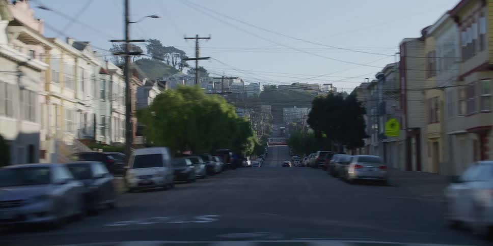 Cars drive along street passing apartment buildings and a cross street that leads to building topped hill