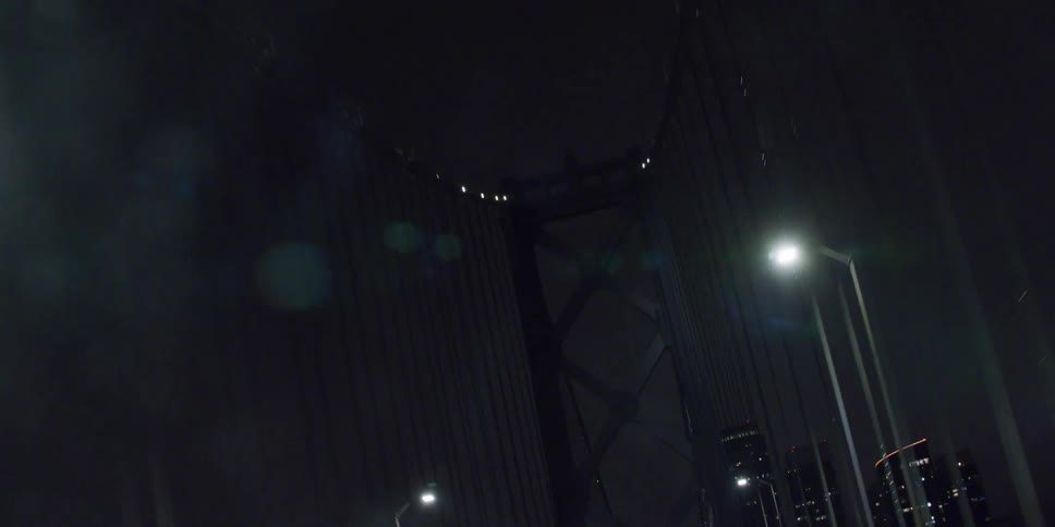 Vehicle crosses suspension bridge leading to city with high rise buildings at night