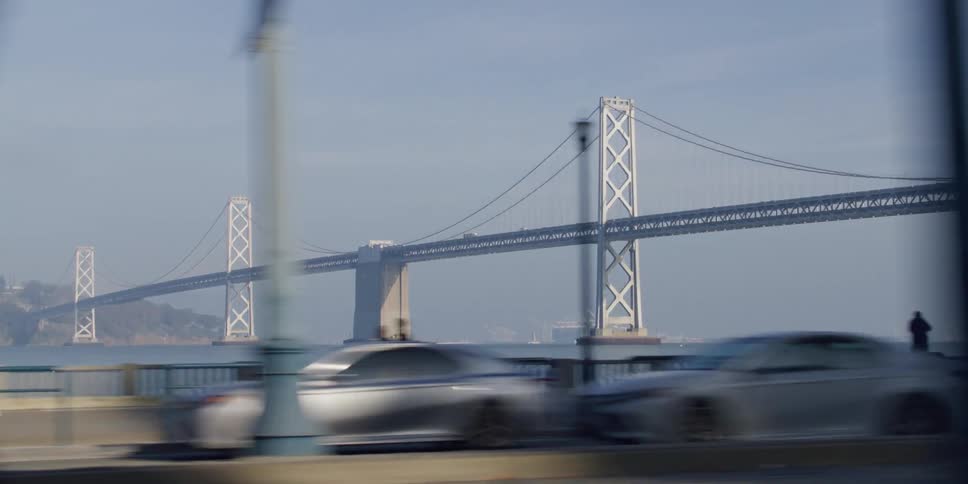 Suspension bridge with suspension cables on vertical suspenders seen from a moving car on highway next to promenade