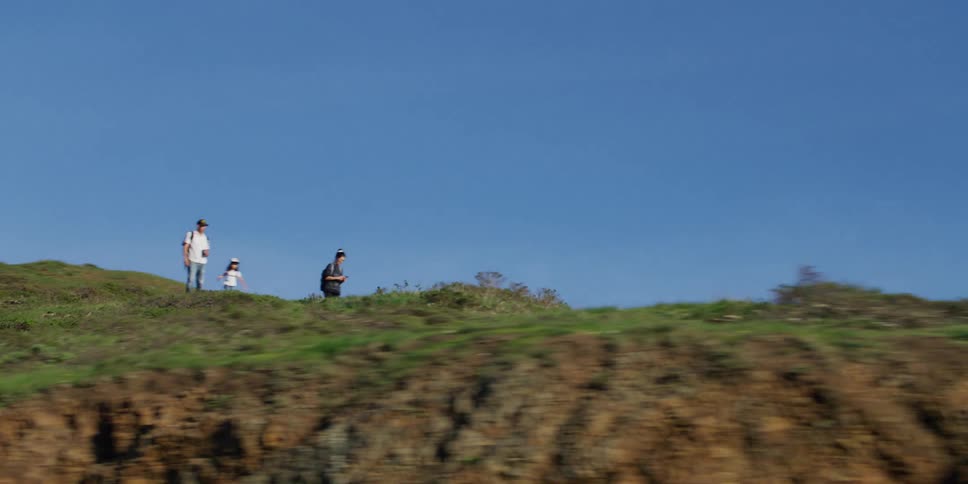 A man follows behind as his son and daughter run along a cliff on the coast