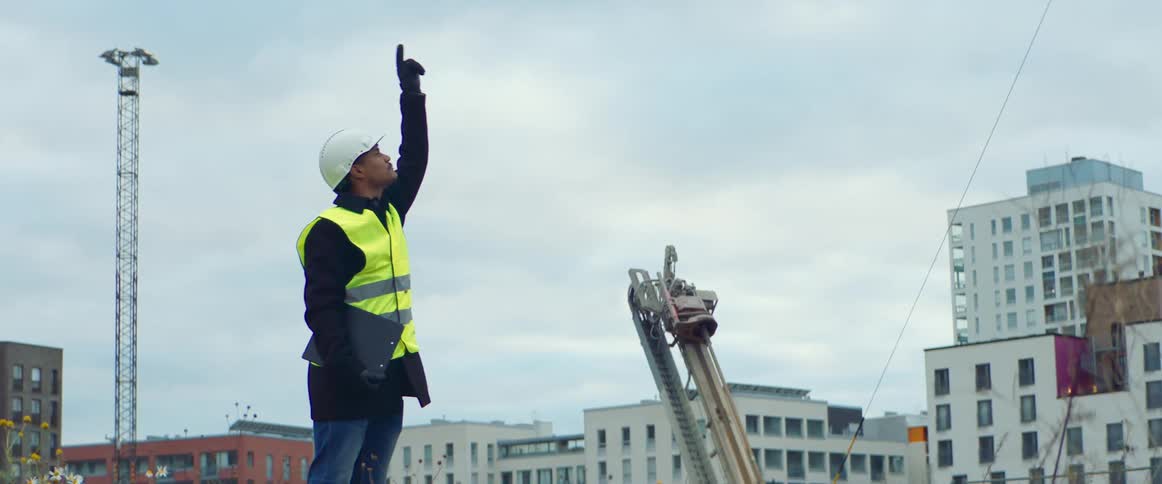 Man wearing a white hardhat and a bright yellow vest supervises a construction site in the city