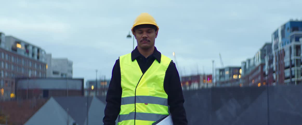 Man wearing a yellow hardhat and a bright yellow vest supervises a construction site in the city