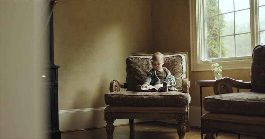 Baby sitting alone at an armchair next to a window leafing through a book