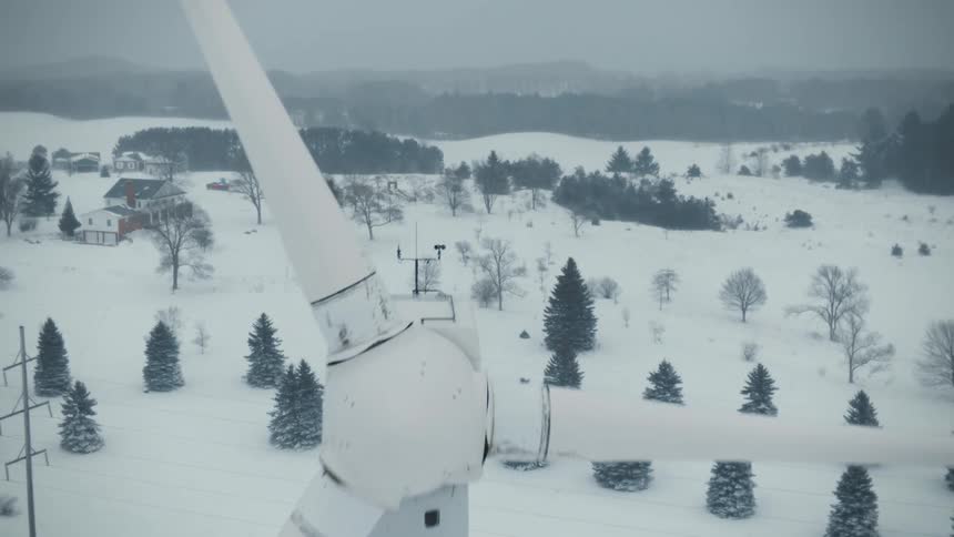 A wind turbine stands with blades still as snow falls on a rural area