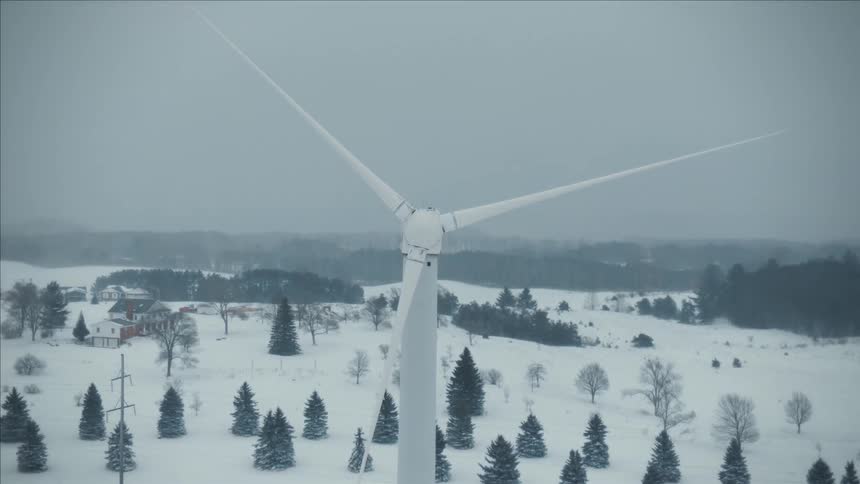 A wind turbine slowly turns above a snow covered ground with trees and forests
