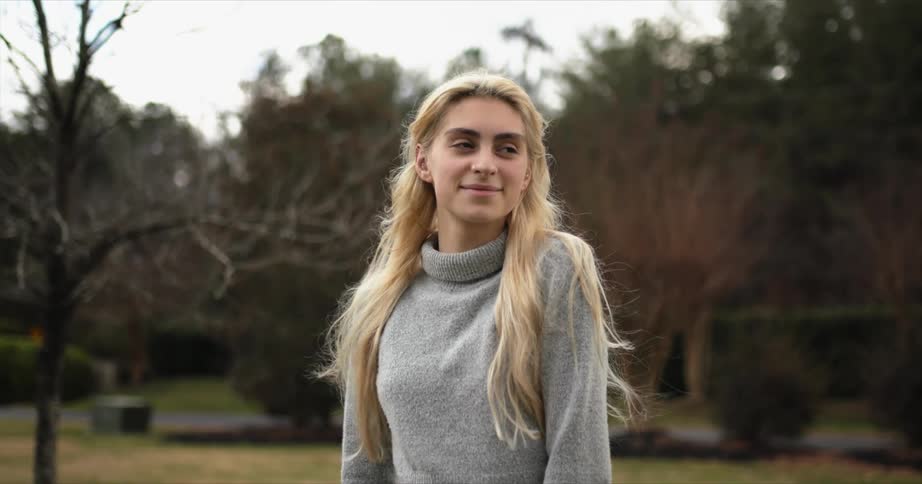 White blonde woman wearing winter clothing standing outdoors next to some trees during fall