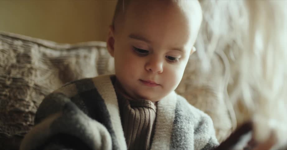 Toddler baby holds smartphone sitting on couch in front of his mother with blonde hair