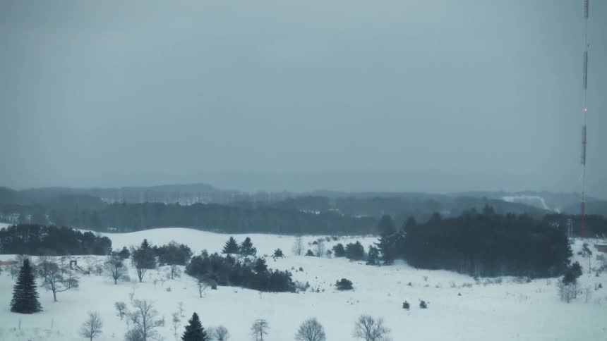 A wind turbine slowly turns above a snow covered ground with trees and forests