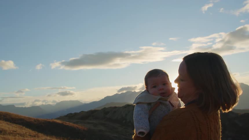 Blonde mother in brown sweater holds and kisses baby on hill at sunset