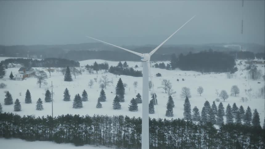 Wind turbine blades slowly rotate near snowy field dotted with conifer trees