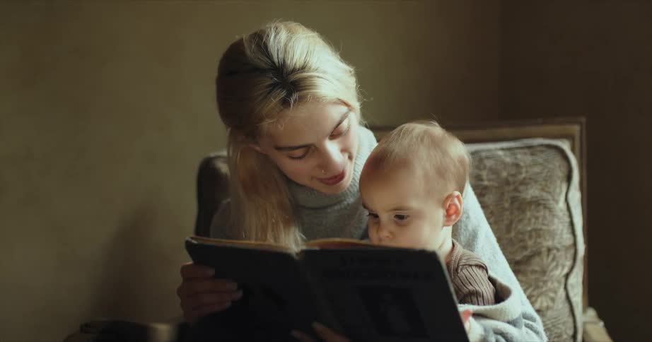 Mother reading a story from a book to her baby sitting on a chair at home