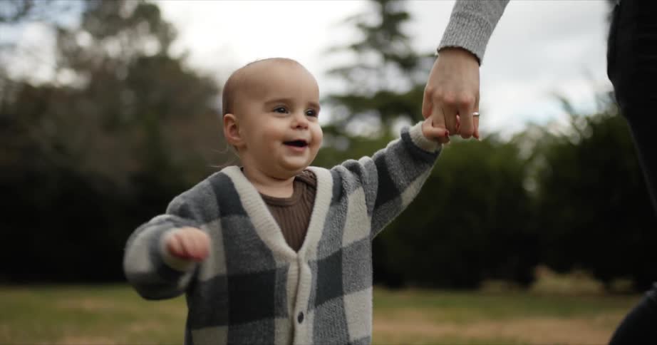 Baby girl running laughing and holding her mothers hand at a grass field next to some trees