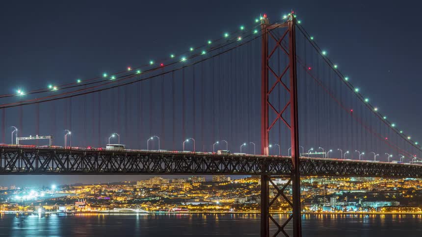 Distant view of vehicles crossing suspension bridge over river with illuminated city buildings in the background