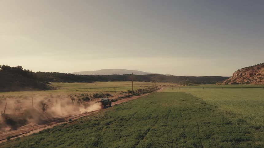 Truck drives on a dirt road leaving a dust trail behind through the fields at morning