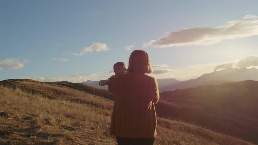 Mother holding a baby in her arms standing on a hill near the mountains at sunset
