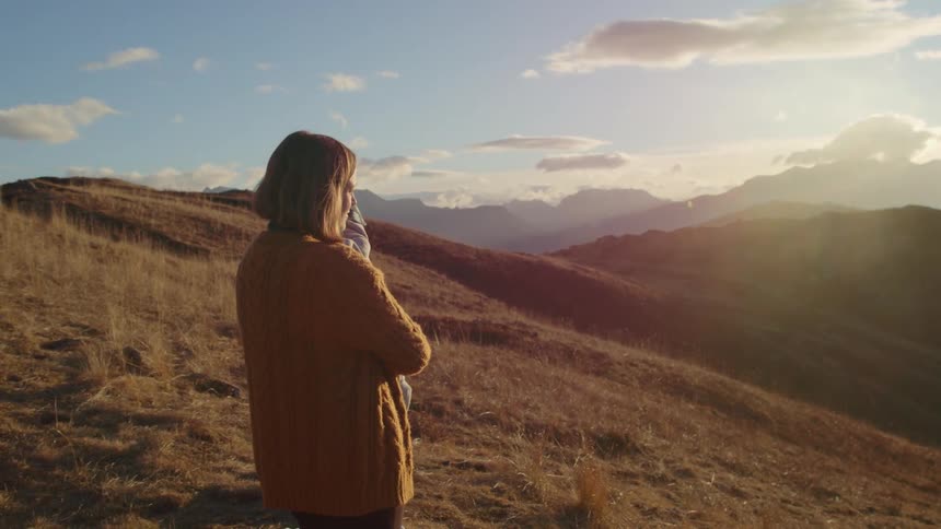 Mother holding a baby in her arms standing on a hill near the mountains at sunset