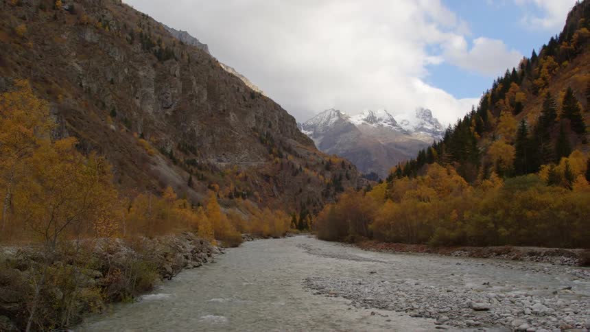 A river runs through a mountain valley lined with trees in fall colors and a snow covered mountain in the distance