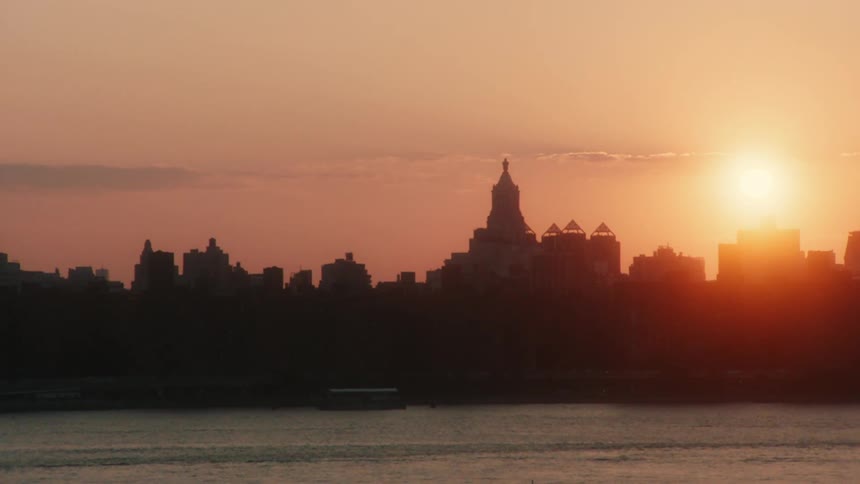 The sun shines brightly in an orange sky behind the silhouette of a city along the coast