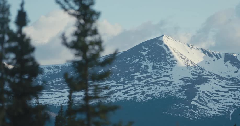 A view through trees as the sun shines on a snow covered mountain with a peak high in a cloudy sky