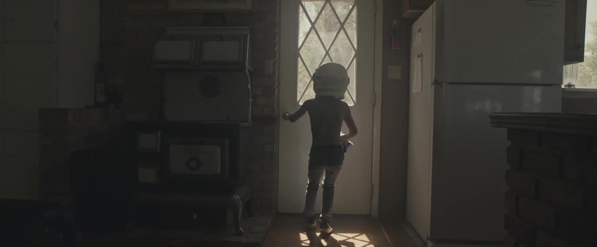 A boy wearing an astronaut helmet and holding a toy shuttle runs to the door and heads outside