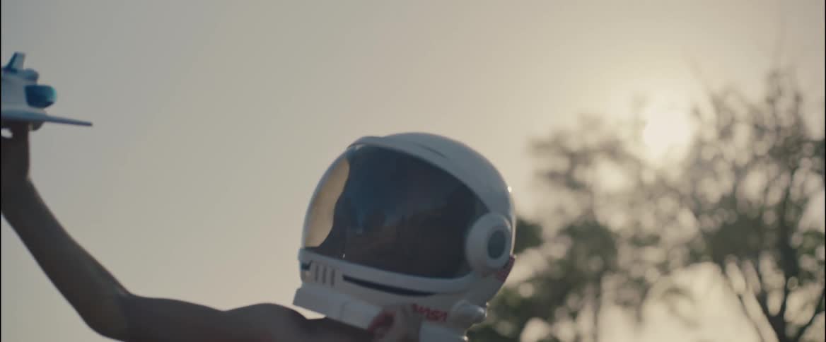 A boy wearing an astronaut helmet holds his toy space shuttle up high as he walks
