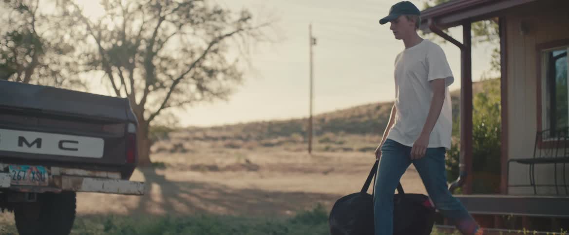 Young man carrying a hand bag walks from a house to a truck parked in the countryside at morning