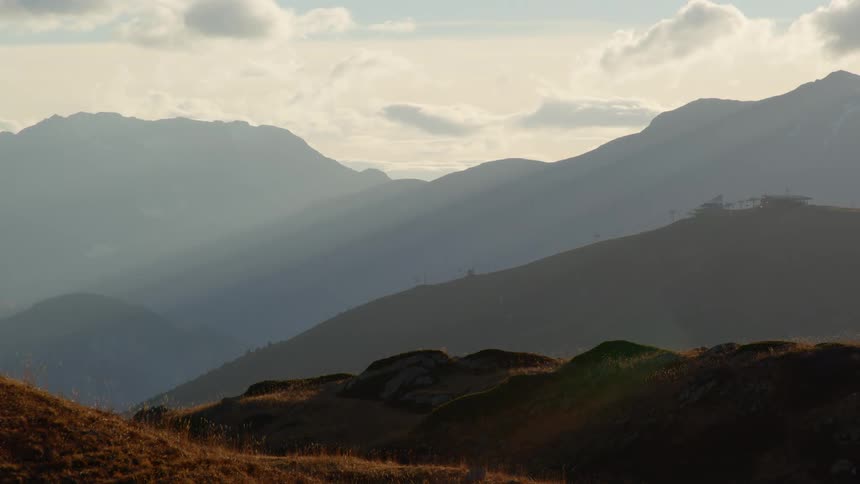 The sun shines on the hills with a mountain range in the distance under a cloudy sky