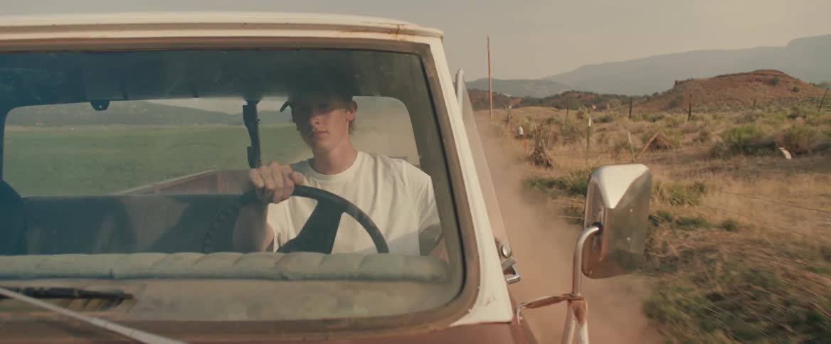 Man drives truck along dirt road passing fenced pasture with misty mountains on the horizon
