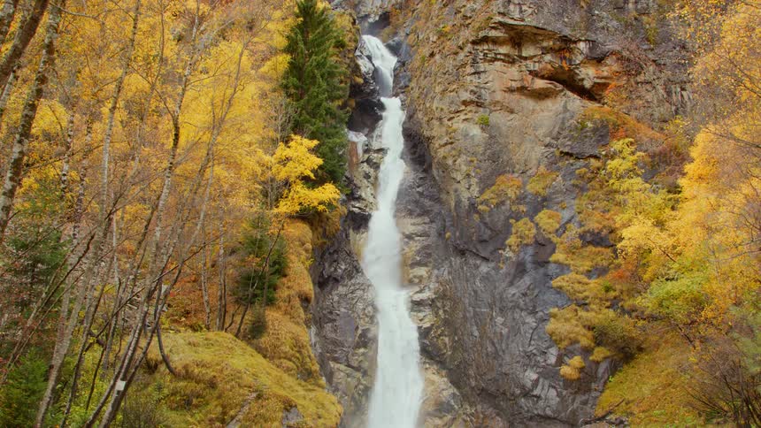 Waterfalls flow down the side of a mountain in a forest of trees with fall colored leaves