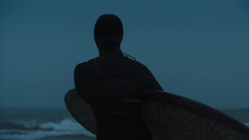 Male surfer in wetsuit holds surfboard standing on beach looking at sea at dusk