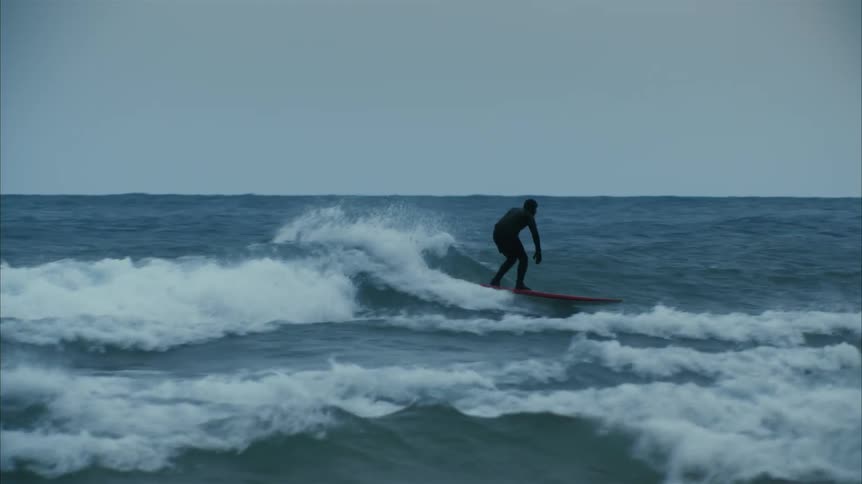 Surfer wearing a full body black wet suit rides a red surfboard on the ocean waves close to the coast