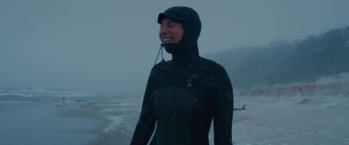 A woman stands on a beach wearing a full wet suit laughing as it snows