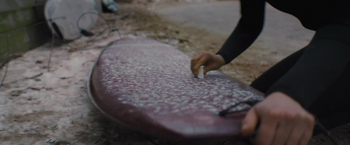 A surfer kneeling rubs wax on her board in the parking lot